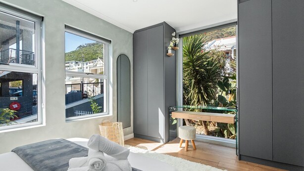 A modern bedroom with a large window, a bed with a white and gray blanket, a wooden bench, and a small table with a basket. - Selbourne Five - Two Bedroom Apartment, Sleeps 4 (Cape Town)