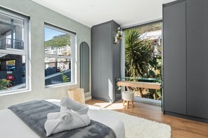 A modern bedroom with a large window, a bed with a white and gray blanket, a small wooden stool, and a glass table with a plant.