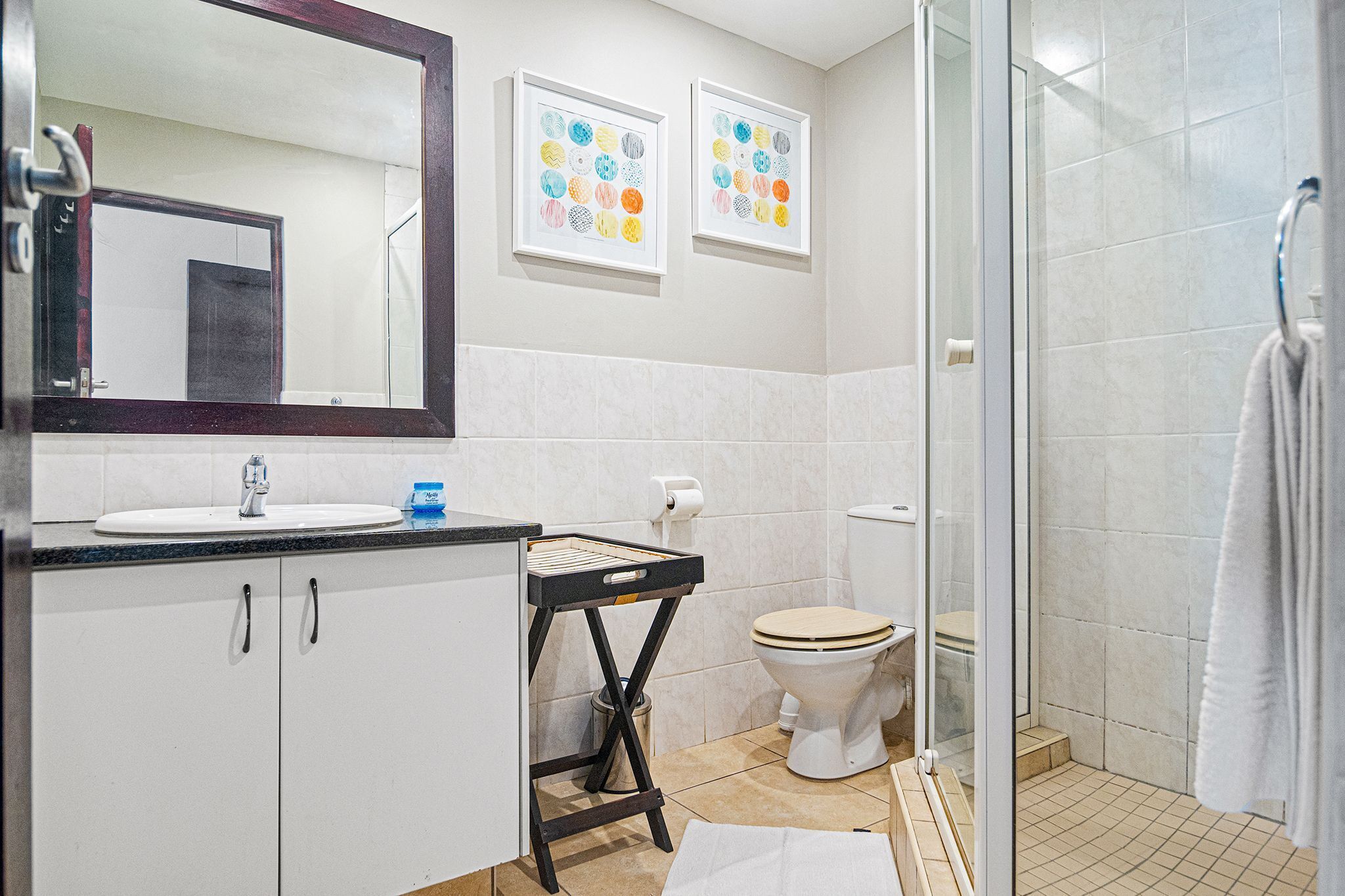 A bathroom with a sink, toilet, and shower. There are framed pictures on the wall and a mirror above the sink.