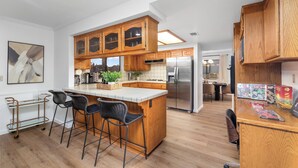 A modern kitchen with wooden cabinets, a stainless steel refrigerator, and a bar area with stools. - Lakeside Retreat Hot Tub, Fire Pit, 4BR/3BA (Victorville)