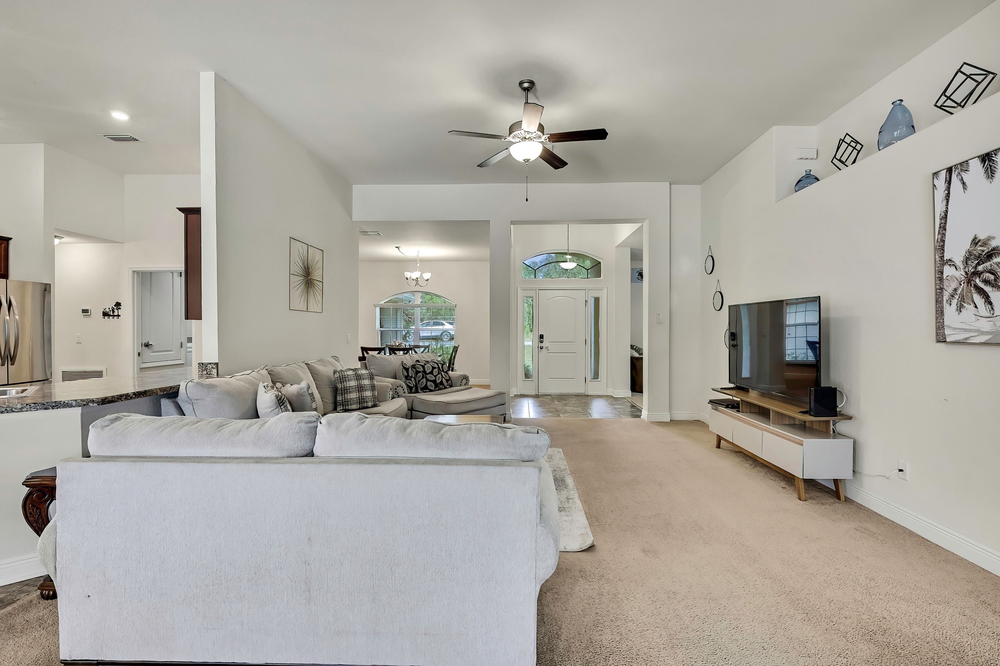A spacious living room with a large sofa, a TV, and a ceiling fan. There is a kitchen area in the background with a dining table and chairs.