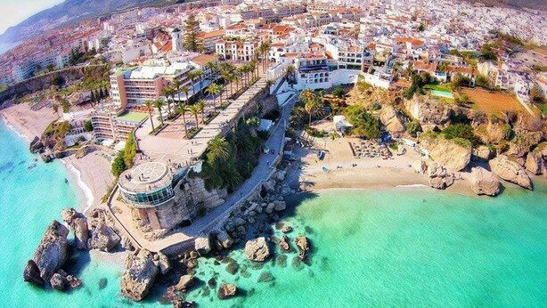 An aerial view of a coastal town with a prominent building featuring a circular terrace and a beach area with clear turquoise waters and rocky outcrops. - Charming Apartment in Burriana With Private Pool! (Nerja)