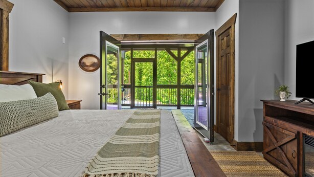 Un dormitorio con una cama grande, vistas a un balcón por puertas abiertas y un televisor sobre un mueble de madera.