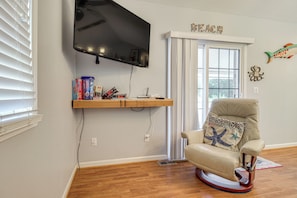 A living room with a mounted TV, a rocking chair, a window with blinds, and a shelf with books and decorative items. - 3 Mi to ICW: Coastal Carolina Home w/ Pool Access (Hampstead)