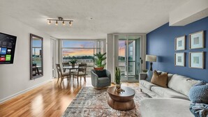 A modern living room with a large TV, a sofa, a coffee table, and a view of the city at sunset.