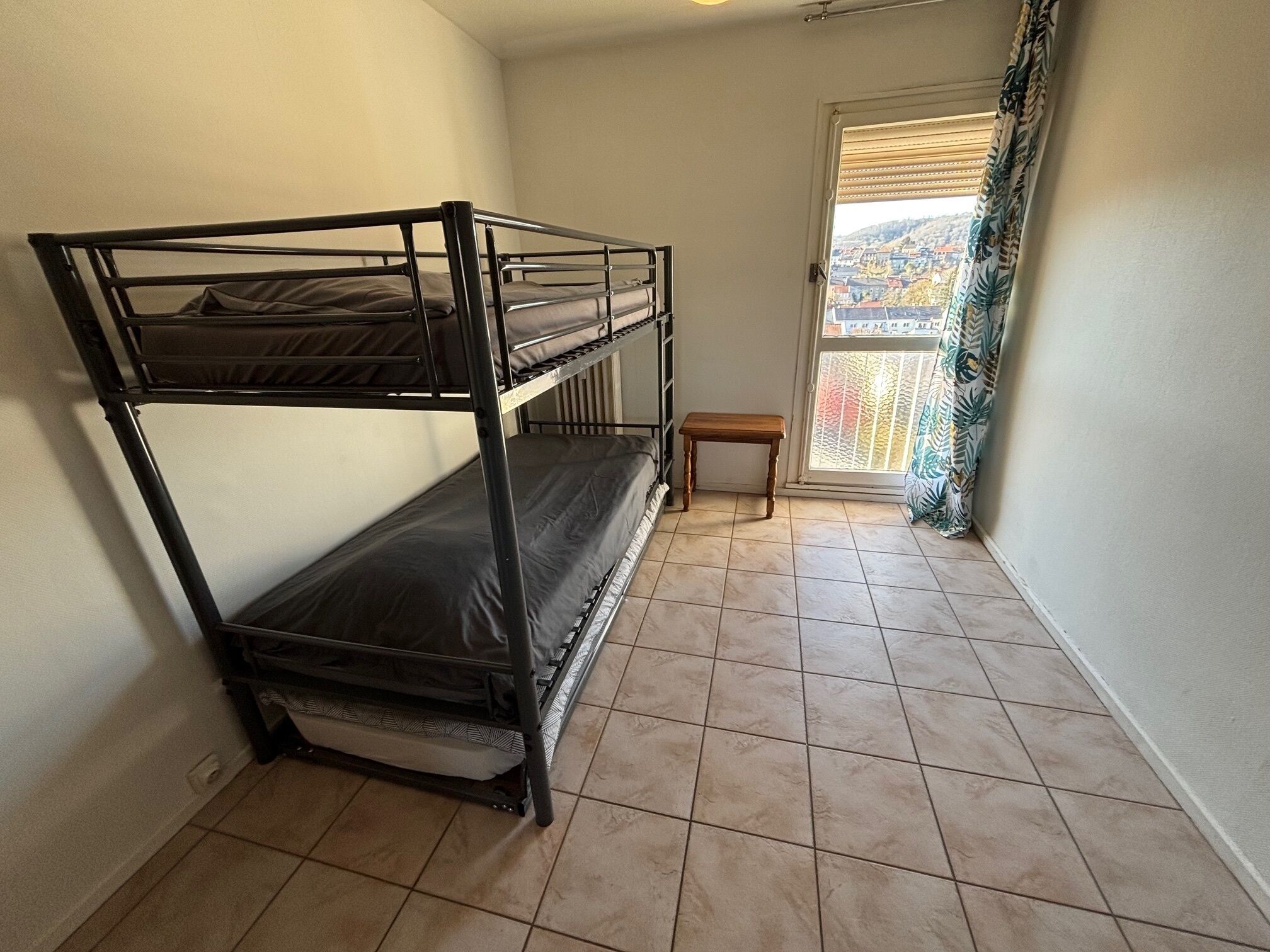 A room with a bunk bed, a small wooden bench, and a view through a window with a curtain.