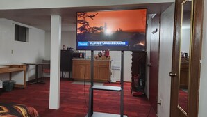 A room with a TV displaying a news broadcast, a wooden desk, a chair, a small kitchenette, and a red carpeted floor.