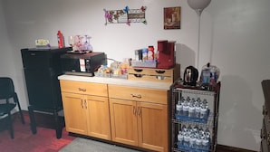 A compact kitchen with a microwave, coffee maker, and various food items on the counter. There is a chair and a small table in the corner.
