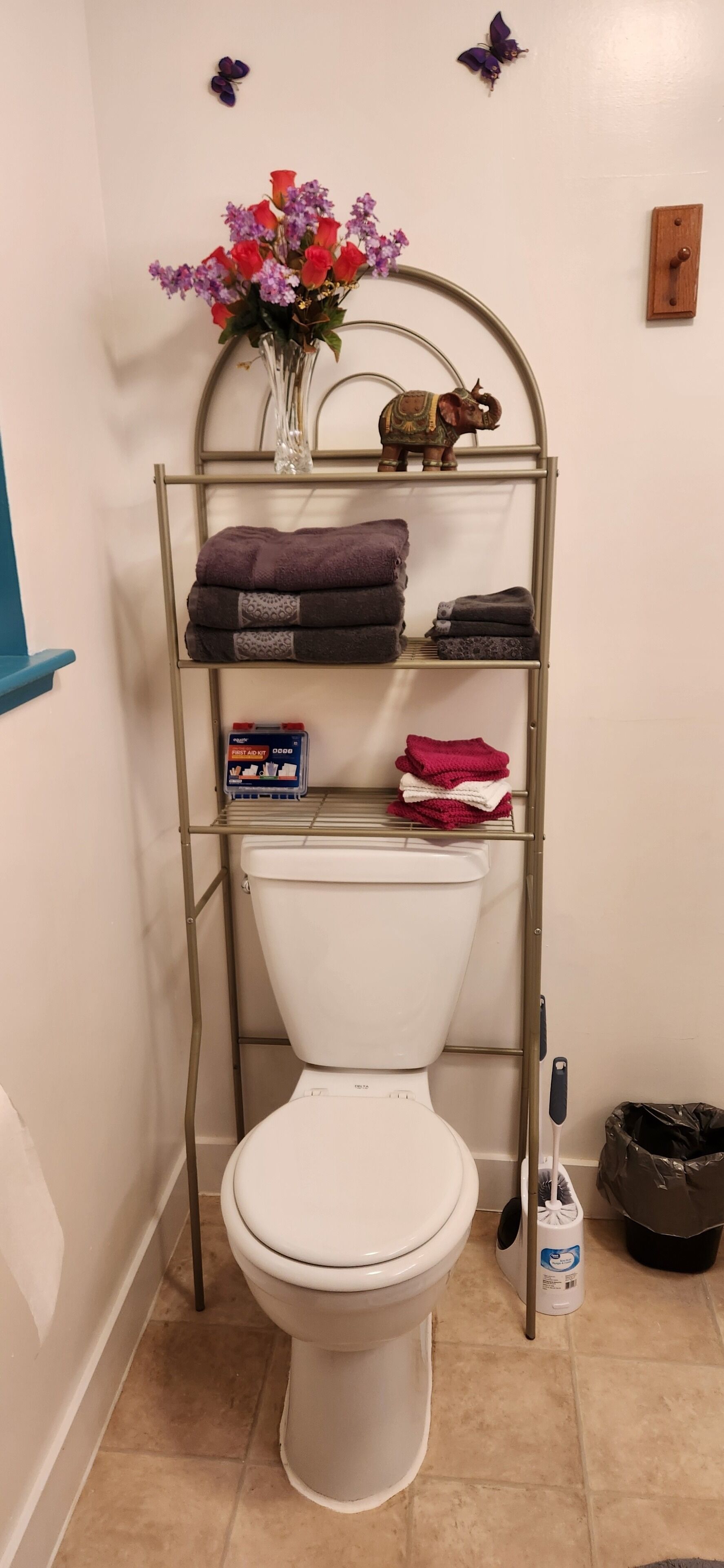 A bathroom with a toilet, a metal shelf with towels and toiletries, a vase with flowers, and a butterfly decoration on the wall.