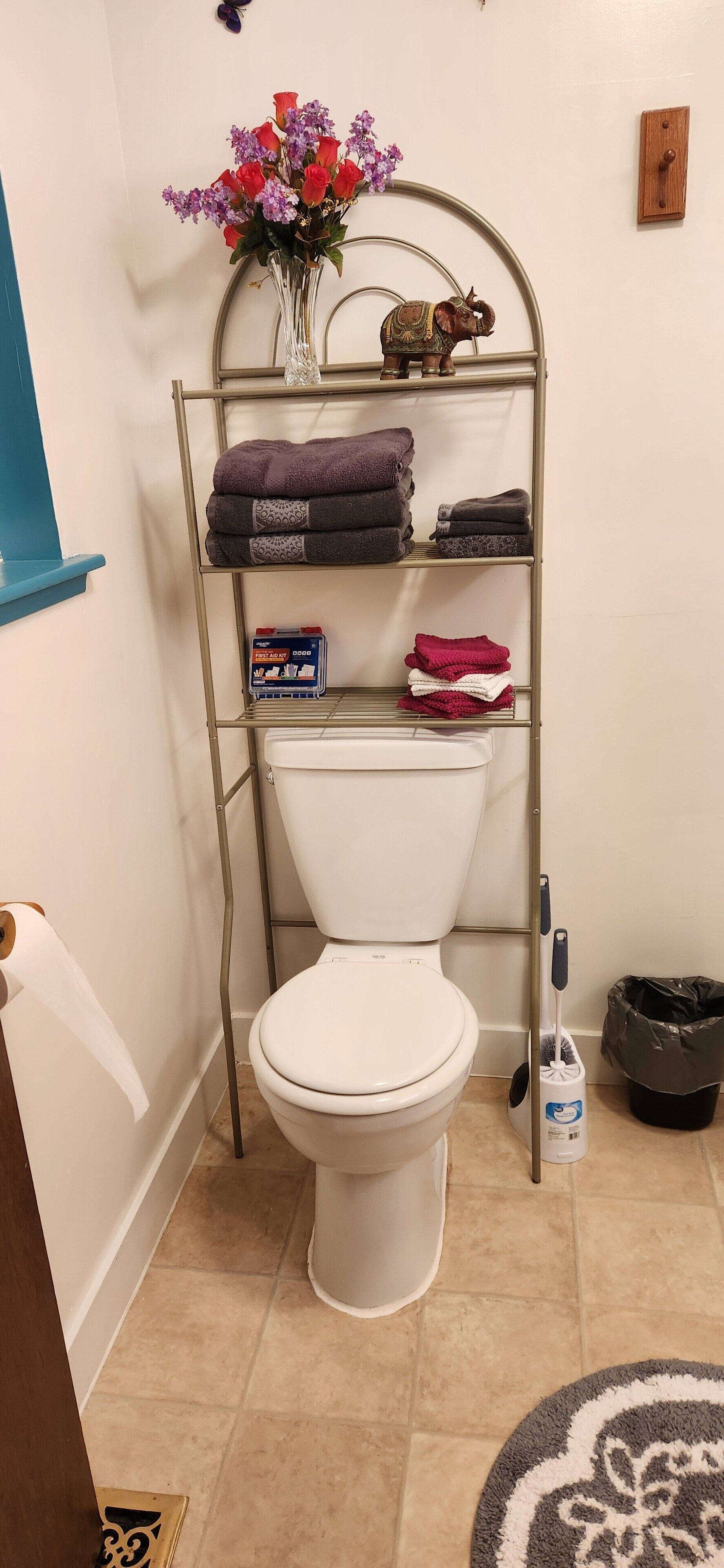 A bathroom with a toilet, a metal shelf with towels, a vase with flowers, and a trash bin.