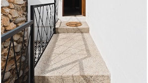 Una escalera conduce a una puerta de madera con un tejado de paja, rodeada de muros blancos y un muro de piedra en un lado.