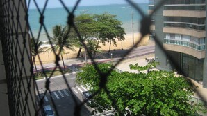 A view through a chain-link fence of a beach with palm trees, a road, and a high-rise building. - 4D5 Your Beach Retreat on the Coast Awaits You 1-minute-from-the-sea  (Vila Velha)