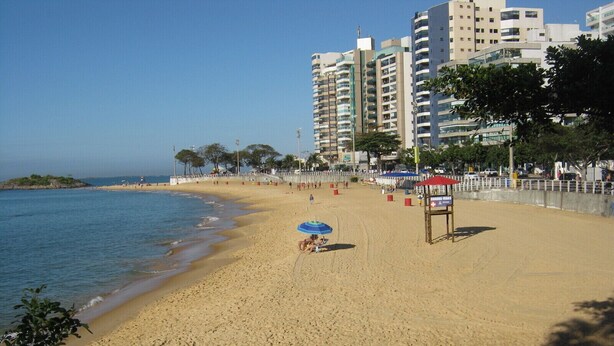 A beach with a lifeguard station, clear skies, and high-rise buildings in the background. - 4D5 Your Beach Retreat on the Coast Awaits You 1-minute-from-the-sea  (Vila Velha)