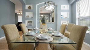 Un comedor con una mesa de vidrio rodeada de sillas tapizadas en tono beige, una lámpara colgante y cuadros en las paredes.