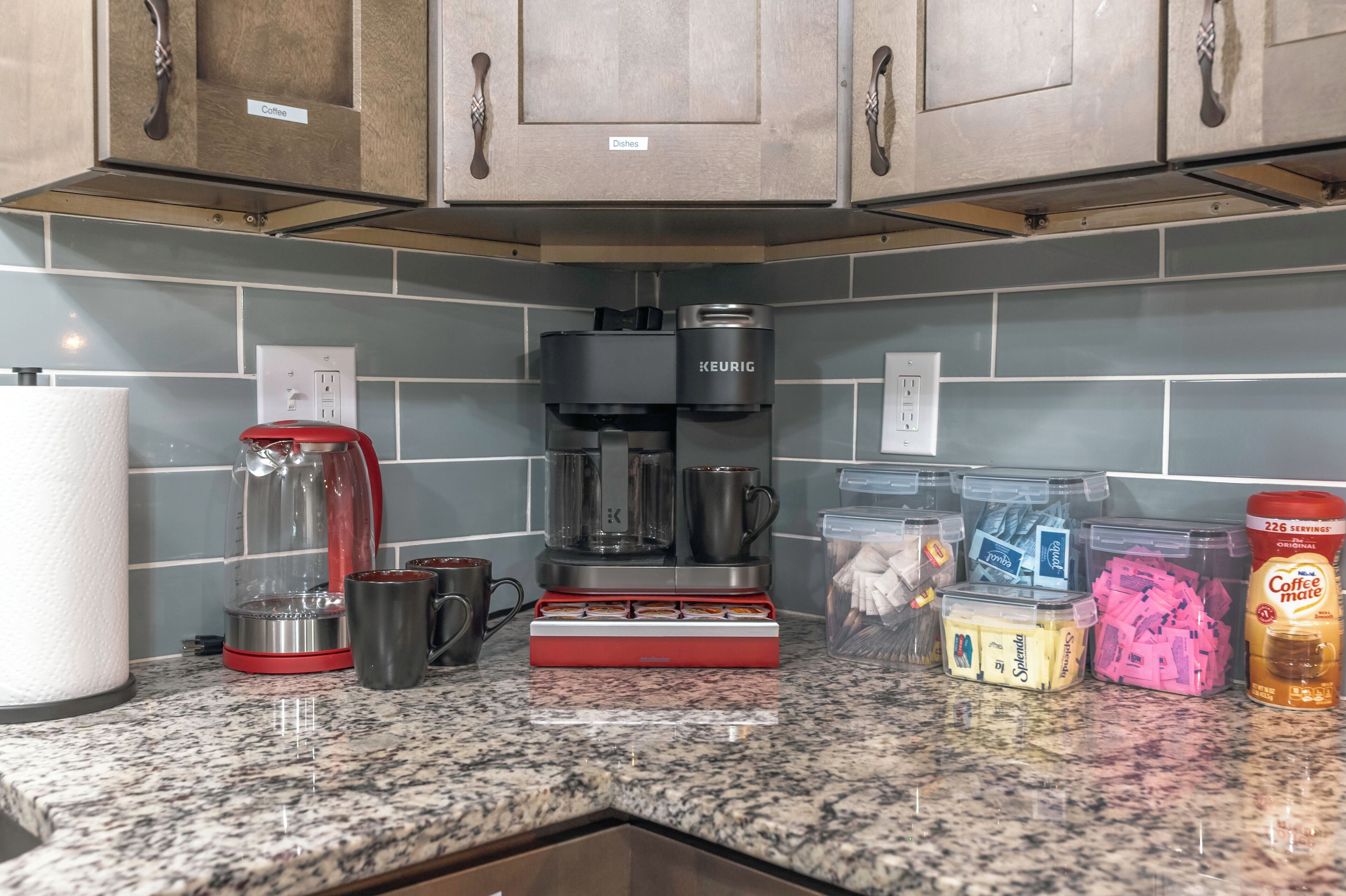 Una cocina con cafetera, hervidor y varios tipos de cápsulas de café sobre la encimera.