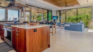 A modern kitchen with a large island, wooden cabinetry, and a built-in oven. There is a dining area with a sofa and chairs, and large windows offering a view of greenery outside. - Villa Enlightened/Ocean View/Walk to Beach (Nosara)