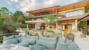 A modern two-story house with outdoor seating, large windows, and a balcony. - Villa Enlightened/Ocean View/Walk to Beach (Nosara)