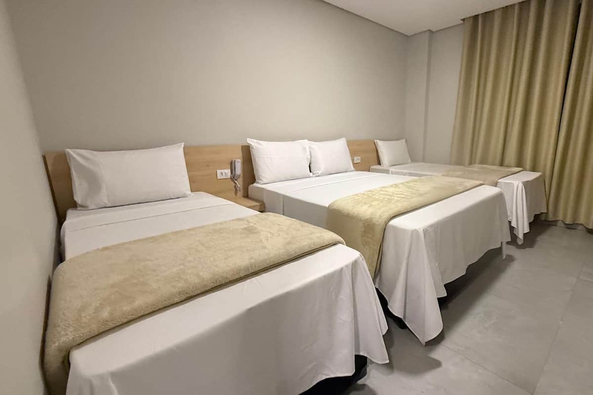 Ein Hotelzimmer mit drei Einzelbetten, jeweils mit weißer Bettdecke und beiger Tagesdecke. Das Zimmer hat eine neutrale Farbgestaltung mit beigen Vorhängen und einem hellen Boden.
