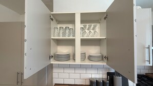 Cozinha com armários brancos, painel de azulejos e bancada de madeira. Há copos e pratos nas prateleiras, e três recipientes rotulados com açúcar, chá e café.