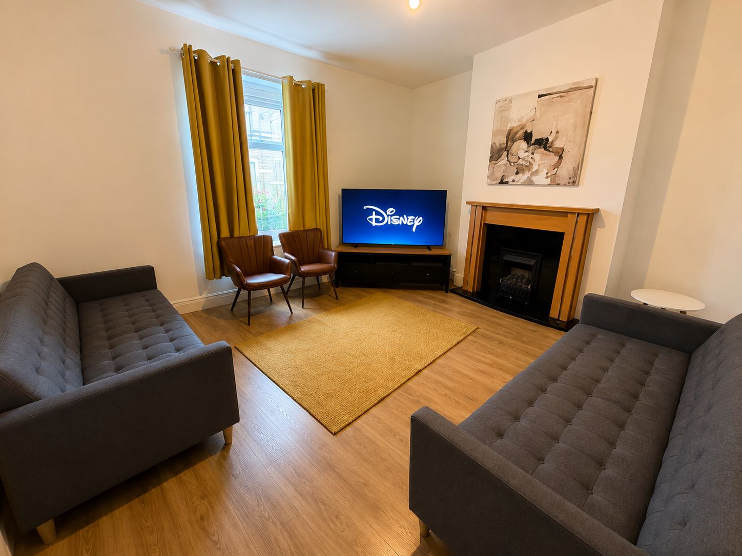 Un salon avec un téléviseur à écran plat affichant le logo Disney, deux fauteuils en cuir, un canapé, une cheminée et un tableau accroché au mur.