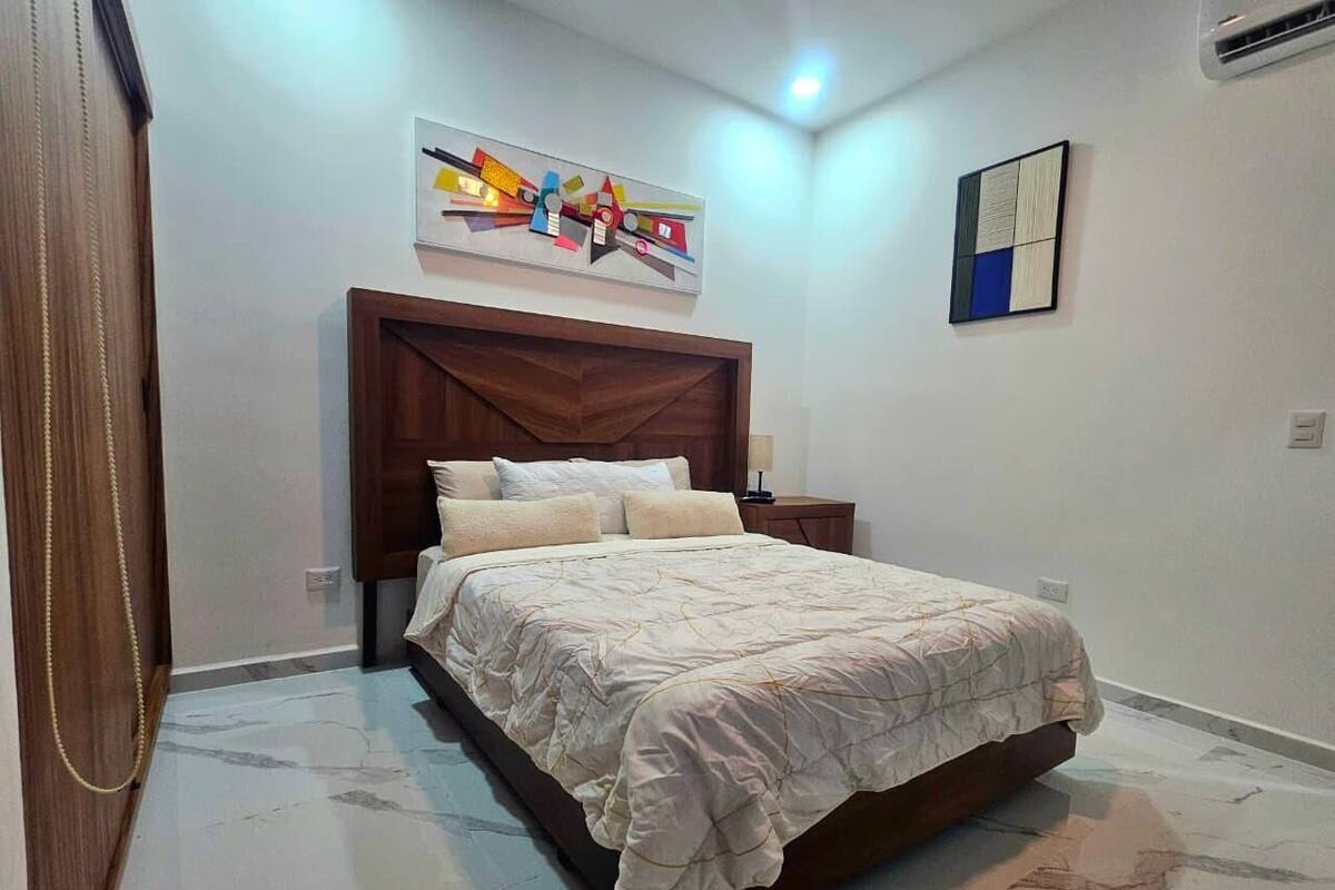 A bedroom with a bed, a wooden headboard, a wall-mounted abstract art piece, a small bedside table, and a wall-mounted light fixture.