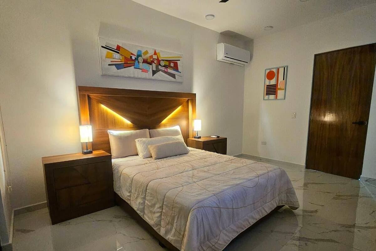 A bedroom with a bed, nightstands, a headboard, wall art, and an air conditioning unit.