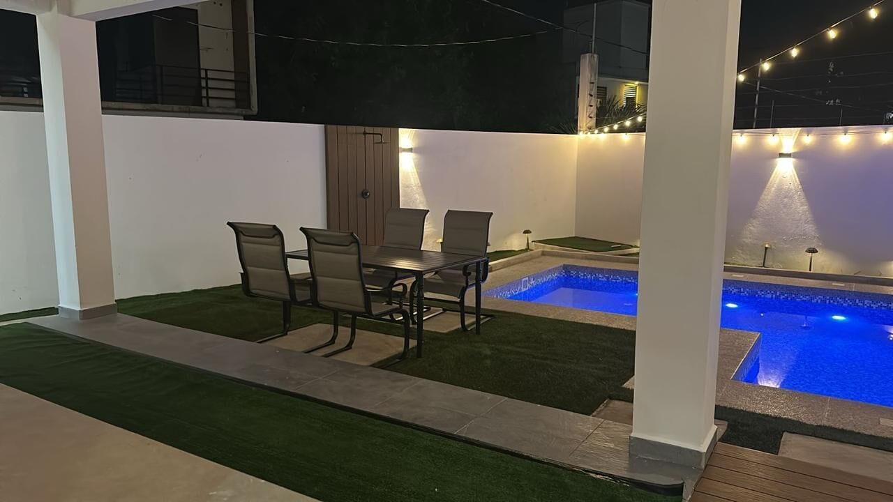 An outdoor patio with a pool, a table with chairs, and string lights.
