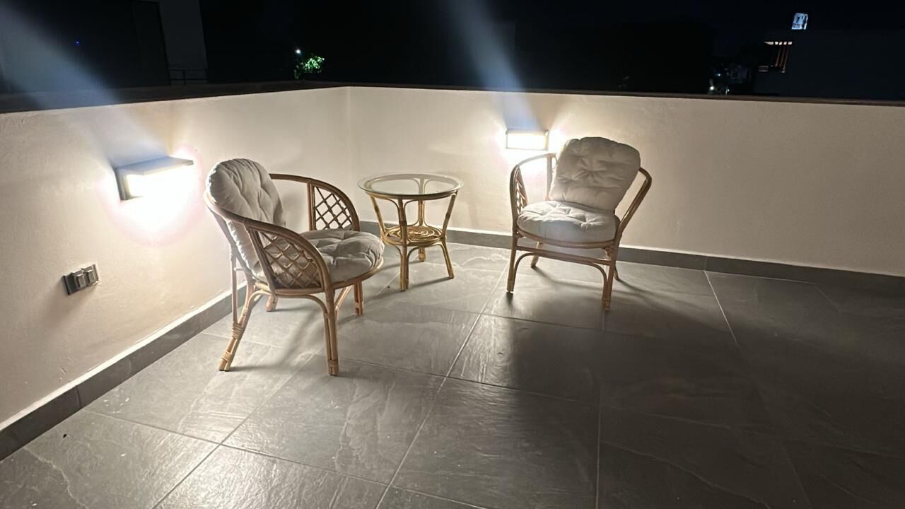 A rooftop terrace with two wicker chairs and a small round table, illuminated by wall-mounted lights.