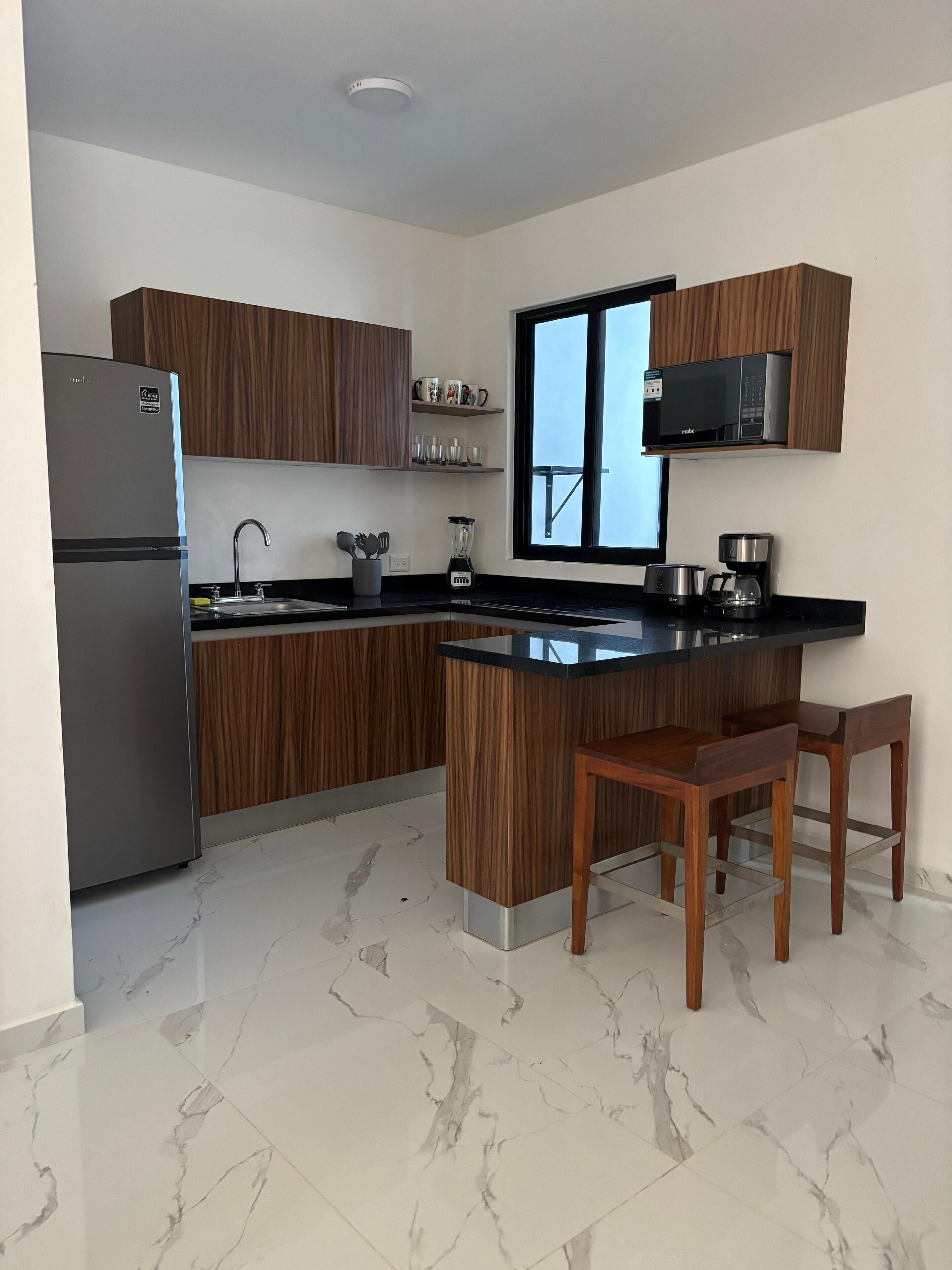 Una cocina moderna con armarios de madera, microondas integrado, refrigerador, pileta y piso de losetas de mármol.