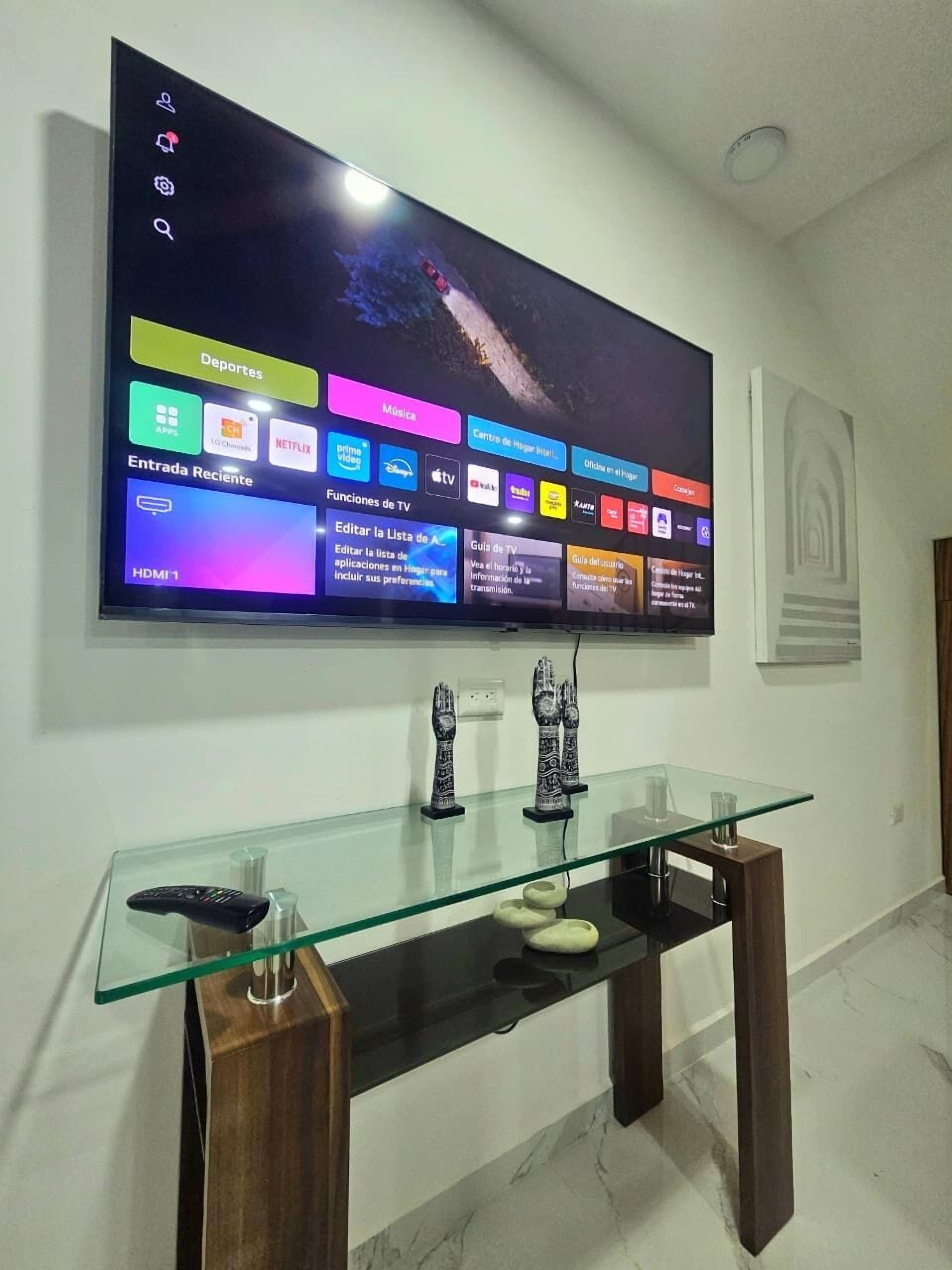 A modern living room with a flat-screen TV mounted on the wall, a glass coffee table with decorative items, and a wooden side table.