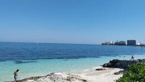 Una playa con agua azul cristalina, costa rocosa y edificios al fondo.