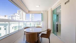 Un balcón con una mesa redonda y dos sillas, una puerta de vidrio que da al baño, y vistas de edificios de la ciudad.