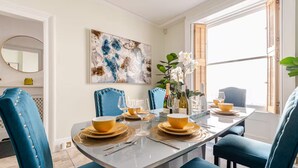 A dining area with a long table set for four, blue upholstered chairs, a large abstract painting on the wall, and a window with wooden shutters.