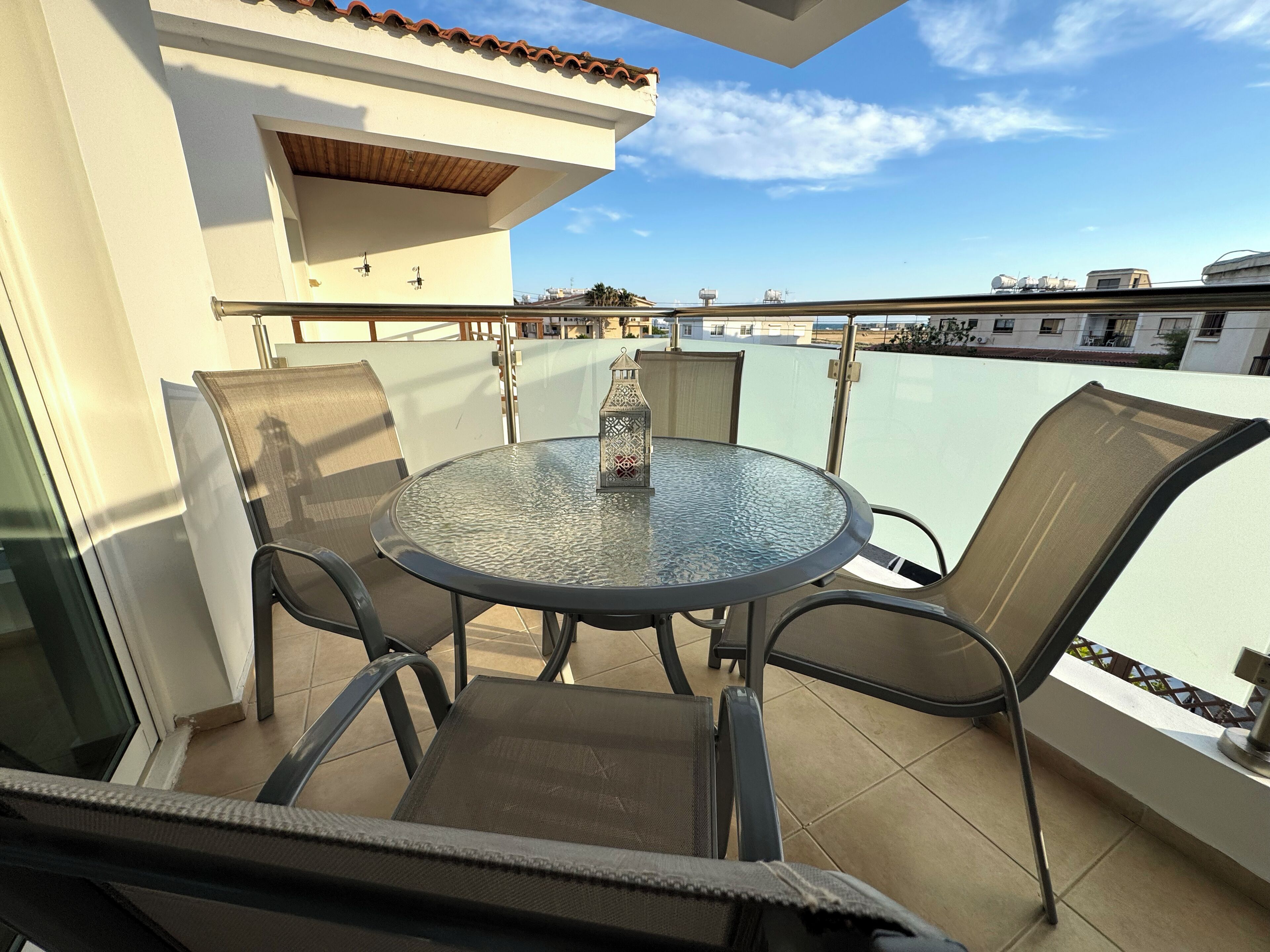 A round glass table with four chairs on a balcony with a view of buildings and a clear sky.