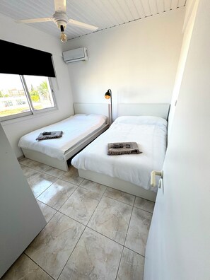 Ein kleines, sauberes Zimmer mit zwei Einzelbetten, einem Deckenventilator, einer Klimaanlage und einem Fenster mit Blick auf umliegende Gebäude.