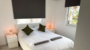 Ein ordentlich bezogenes Bett mit zwei Kissen, einer Nachttischlampe und einem aufgerollten Handtuch auf einer weißen Bettdecke.