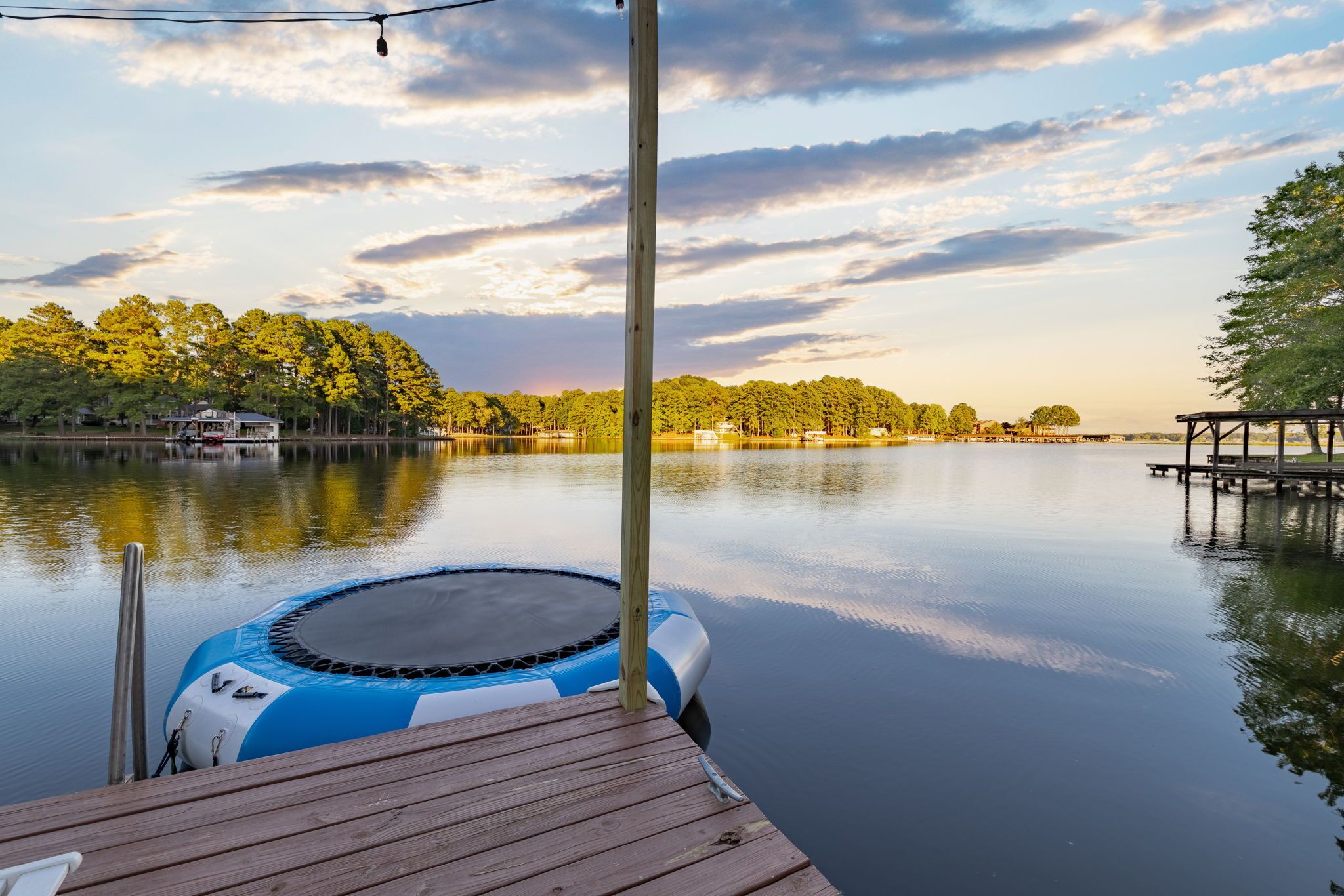A tranquil lakeside setting with a wooden dock, a blue and white inflatable pool, and a clear view of the surrounding trees and distant buildings.