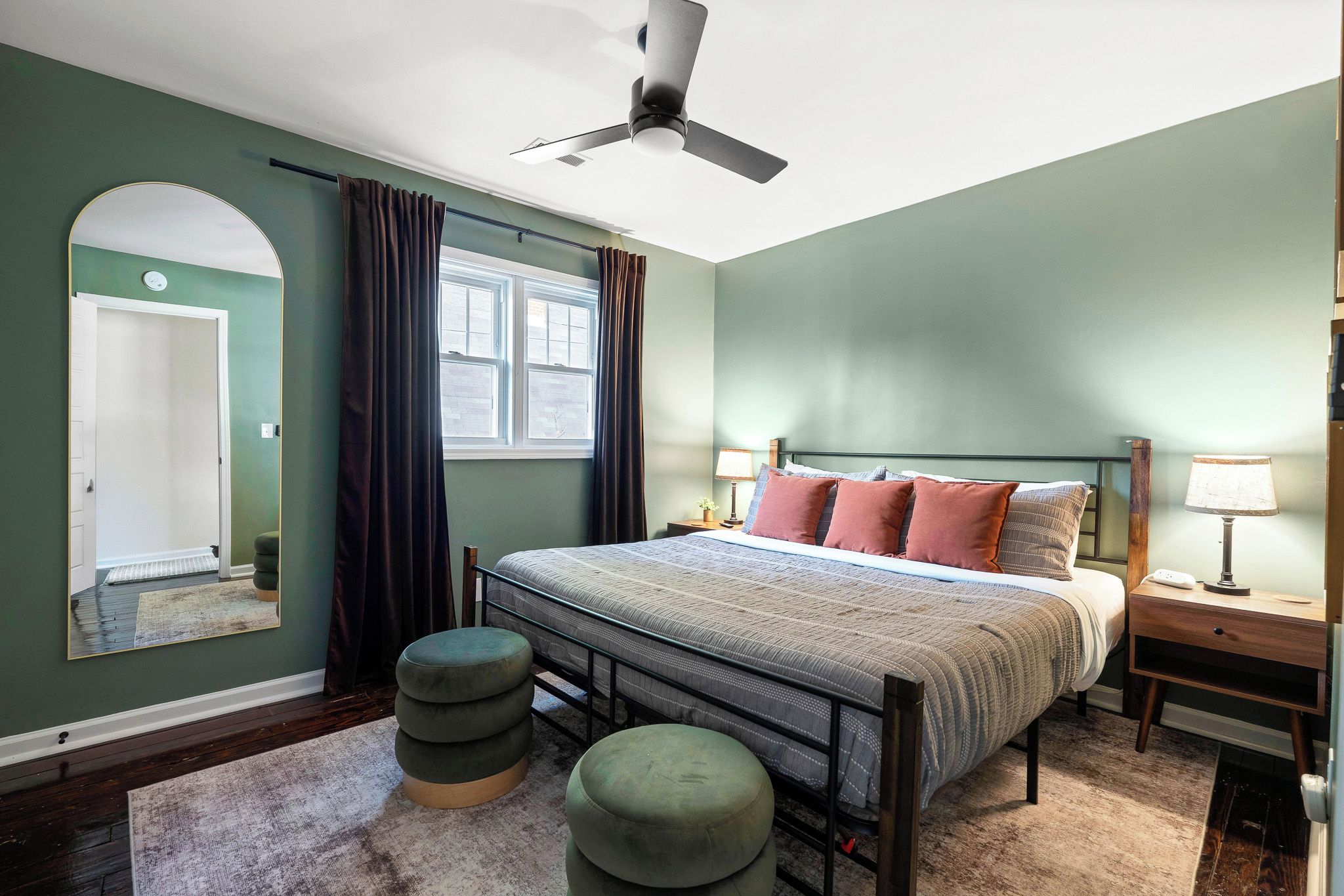 Une chambre à coucher comprenant un lit, des tables de chevet, un ventilateur de plafond, une fenêtre avec des rideaux, un miroir et une porte.