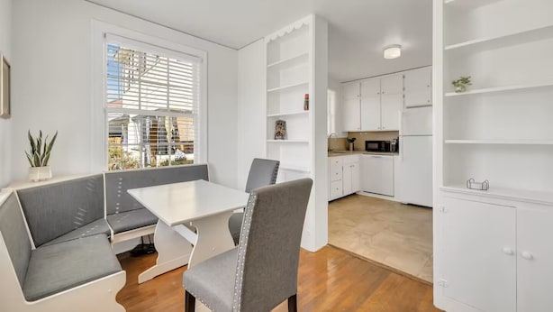 Eine moderne Küche mit weißen Schränken, ein Essbereich mit einem weißen Tisch und grauen Stühlen und eine eingebaute Bank mit grauen Kissen.