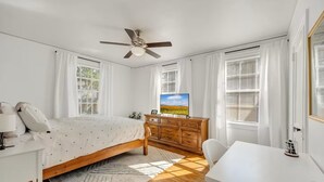 Ein Schlafzimmer mit Bett, Schreibtisch, Kommode, Deckenventilator und großen Fenstern mit Jalousien.