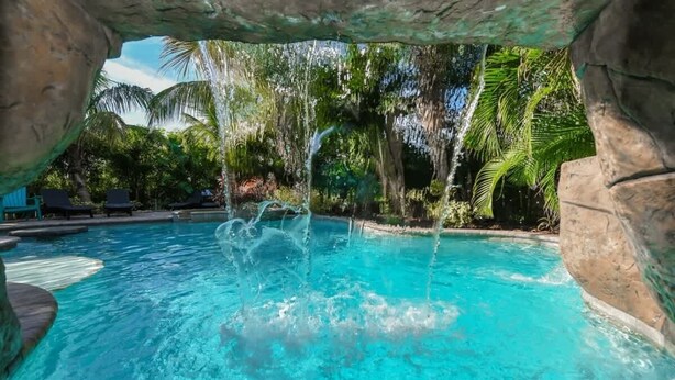Ein klares Schwimmbecken mit Wasserfall, umgeben von Palmen und Liegestühlen.