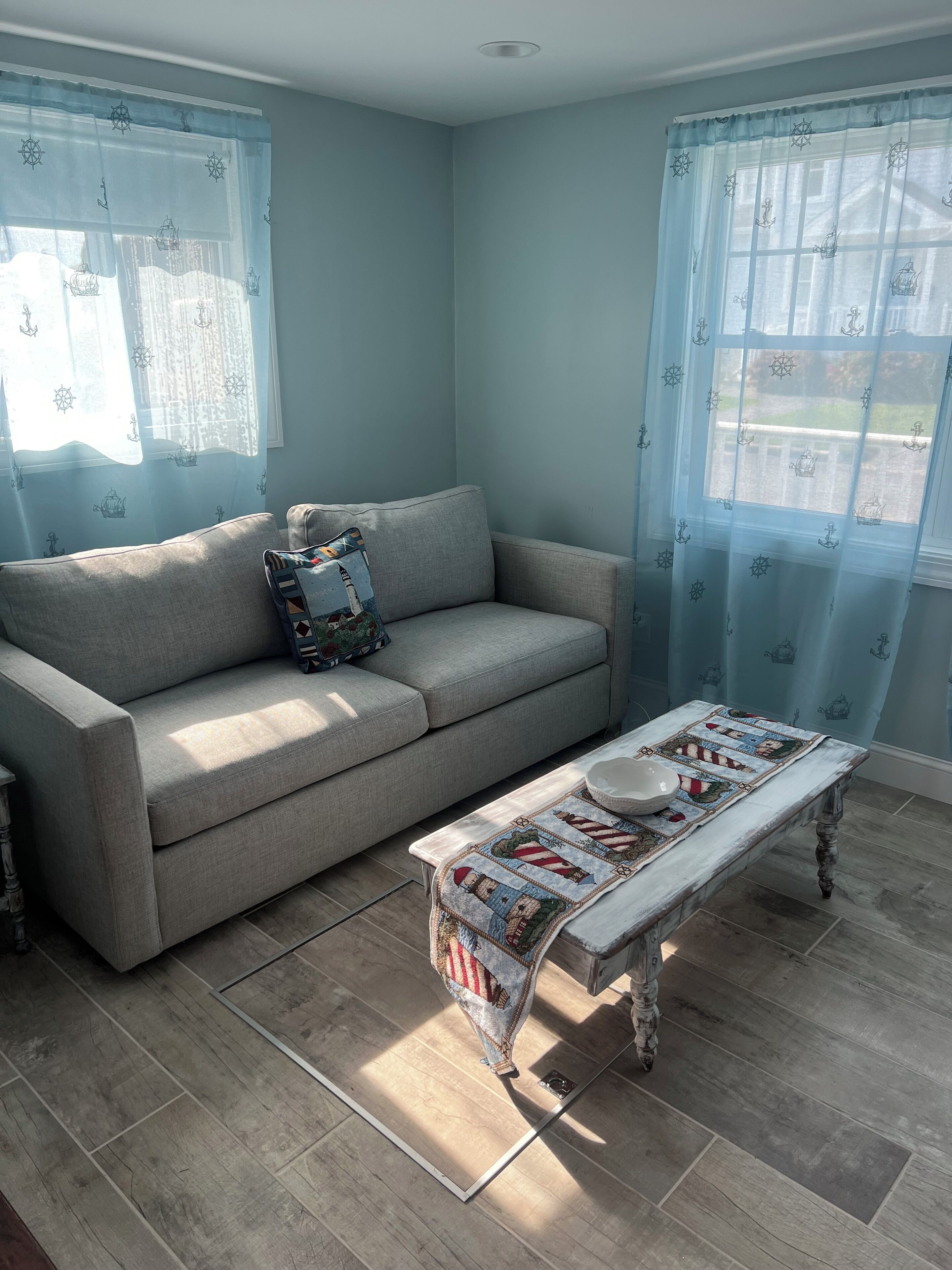 A living room with a grey sofa, a wooden coffee table with a quilt, and light blue curtains with anchor patterns.