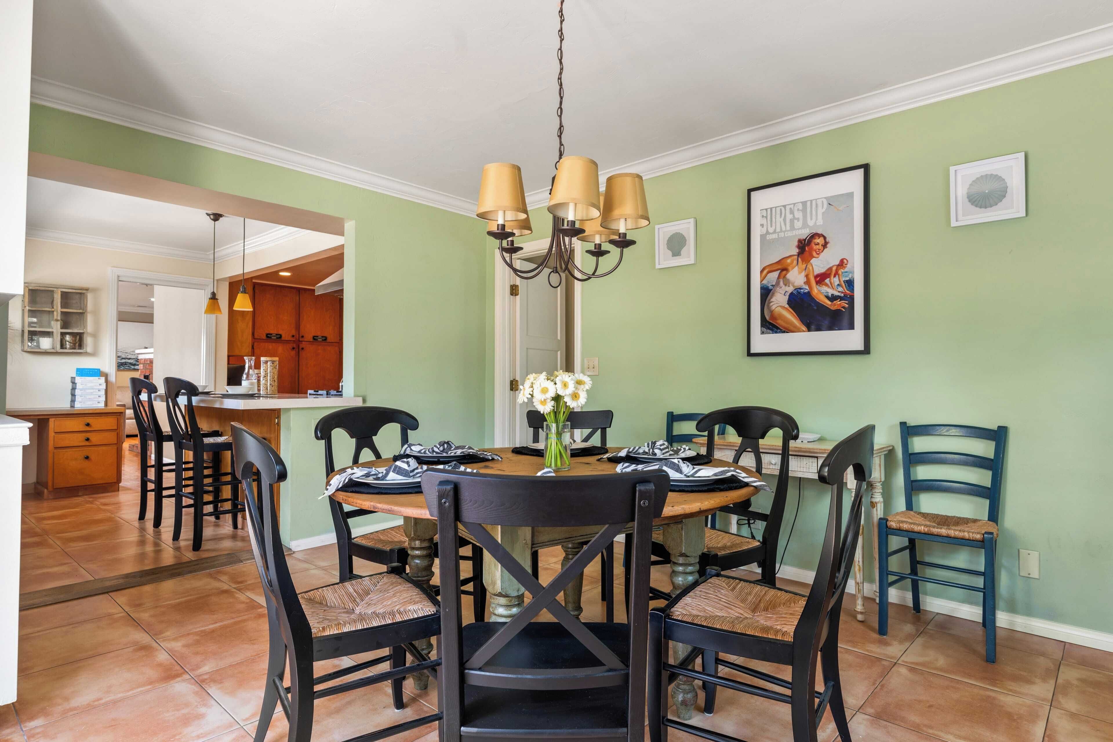 Een eetruimte met een ronde tafel, zes stoelen en een kroonluchter. Achtergrond is een keuken met houten kastjes en een aanrecht.