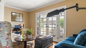 A living room with a blue sofa, a chessboard, a lamp, a desk with a computer, and a large window with a view of the outside. - Point Loma Home w/ Putting Green, Firepit + Game Room & Beach Access Zola Street by Avantstay (San Diego)