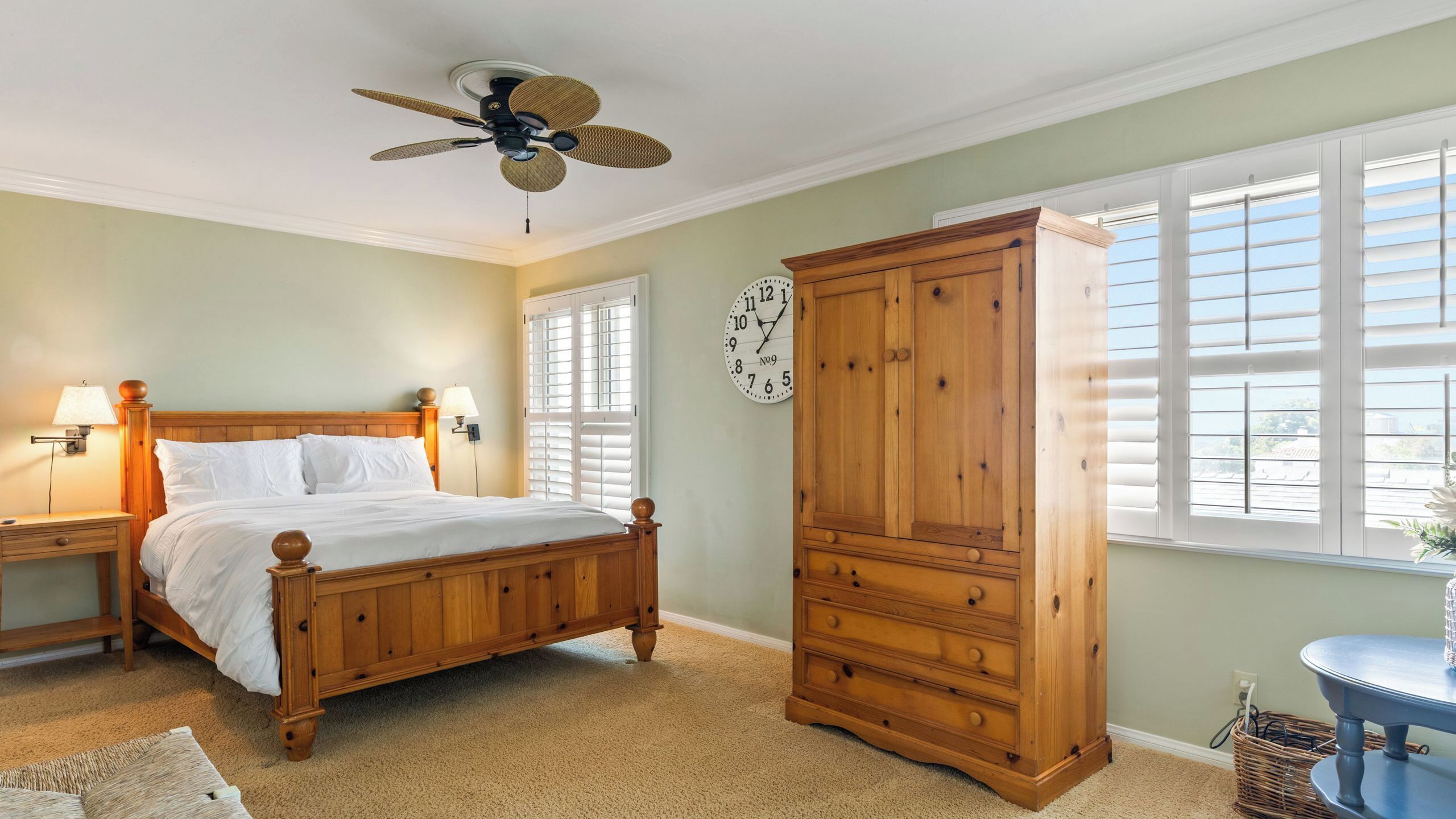 Ein Schlafzimmer mit einem hölzernen Bett, einem Nachttisch, einer Kommode, einer Wanduhr und einem Deckenventilator.