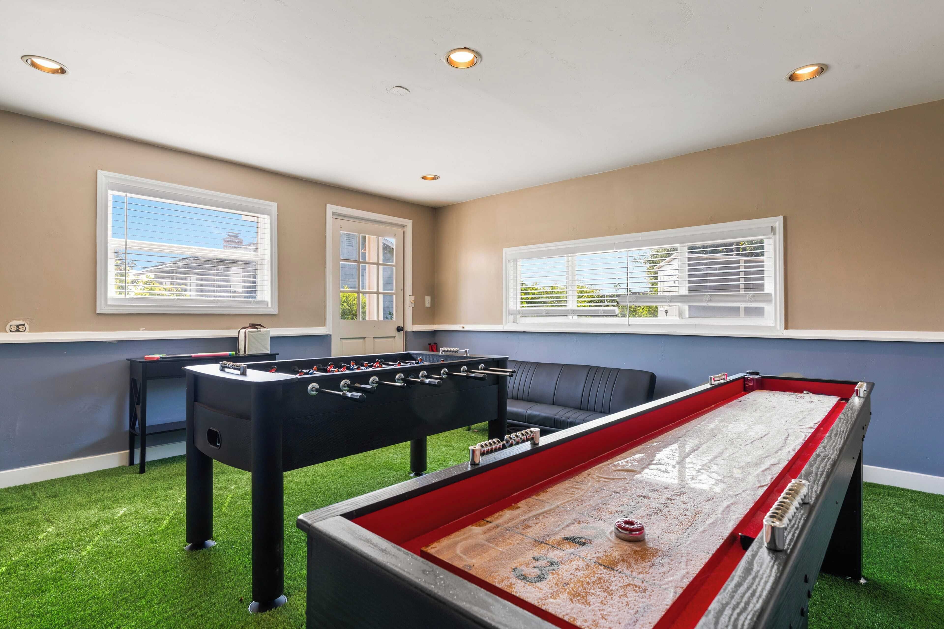 A room with a foosball table, a shuffleboard table, and a sofa. There are windows with blinds, a door, and a ceiling with recessed lighting.