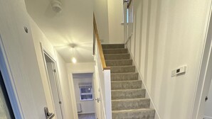 A modern interior with a carpeted staircase, a welcome mat, and a hallway leading to a room with a window. - Modern Detached 4BR House – Super-King En-suite, Garden, Parking, Driveway (Sturry)