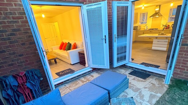A patio with a blue sofa and cushions, a wicker table, and a view into a kitchen and living area through open doors. - Modern Detached 4BR House – Super-King En-suite, Garden, Parking, Driveway (Sturry)