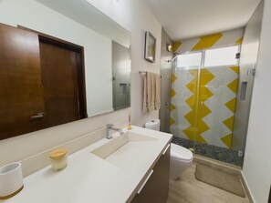 Ein modernes Badezimmer mit einem großen Spiegel, einem weißen Waschbecken, einer Toilette und einer Duschkabine aus Glas mit gelb-weißen Zickzackstreifen.