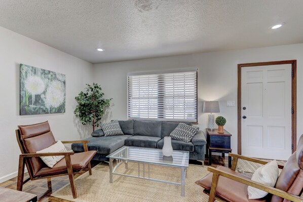 A living room with a sectional sofa, two leather chairs, a glass coffee table, a potted plant, and a painting on the wall.